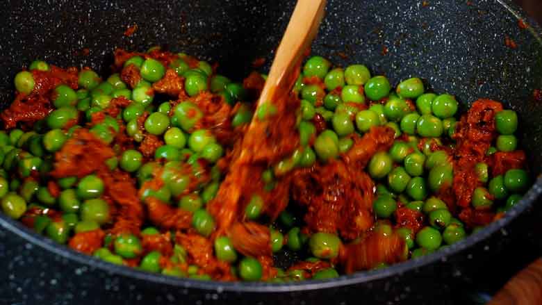 A close up shot of a wooden spatula stirring wet spice paste into fresh green peas in a grey and white speckled non stick pan - Malvani Green Peas and Walnut Curry Recipe
